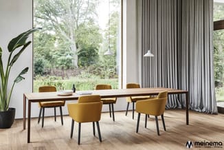 Vergaderruimte met houten tafel, gele stoelen met zwarte poten, groene plant, grote ramen, grijze gordijnen en hanglamp.