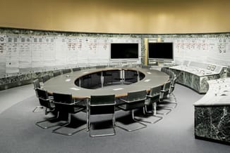 Ronde vergadertafel met grijs blad en houten rand, zwarte ergonomische stoelen, centraal scherm, technische schema's aan wand