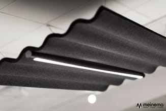 Zwarte akoestische hangende panelen met golvende randen en geïntegreerde lineaire verlichting tegen een wit plafond.