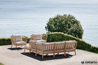 Houten loungeset met grijze kussens op terras aan water, inclusief hoekbank en twee loungefauteuils.