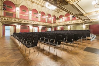 Conferentiezaal met rijen zwarte gestoffeerde stoelen met metalen frame op houten vloer, rode wanden en gouden ornamenten.
