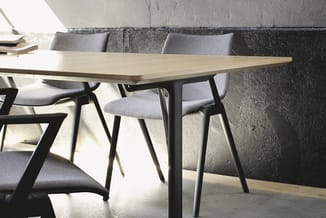 Vergadertafel met licht houten blad en zwart metalen onderstel, omgeven door grijze stoelen in moderne werkomgeving met beton