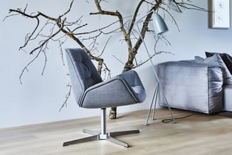 Grijsblauwe draaistoel met metalen voet en armleuningen naast zilveren staande lamp en houten takken tegen witte muur.
