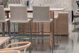 Kantoorruimte met houten tafel, grijze barkrukken met chromen poten en textielwandafwerking op achtergrond.