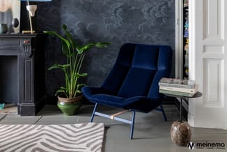 Blauwe fluwelen loungefauteuil met voetenbank in donkere interieurruimte met zwarte schoorsteen, groene plant en boekenplank.