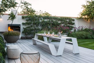Moderne witte picknick/vergadertafel op grijze houten terras met bijzettafels, loungesessel en vuurkorf tegen witte muur met