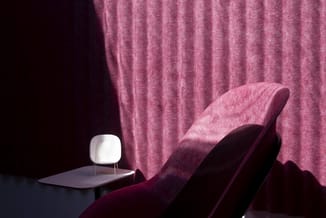 Roze fluwelen fauteuil tegen geplooide gordijn, met witte tafellamp op grijs bijzettafeltje.