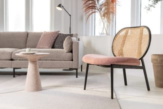 Moderne loungeruimte met beige sofa, roze-bruine fauteuil met ratanrug, ronde travertijnlook tafel en grote ramen.