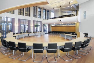 Grote vergaderruimte met ronde houten tafel, zwarte kantelstoelen, houten vloer, grote ramen, gele en witte wanden, tweede ni