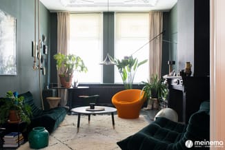 Moderne woonkamer met groene muren, zwarte radiator, oranje fauteul, groene fluwelen sofa, beige vloerkleed, veel kamerplante