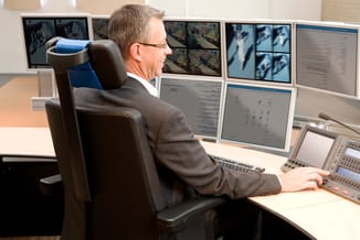 Man in grijsbruine pak zit in zwarte ergonomische bureaustoel aan werkplek met meerdere monitoren en bedieningspaneel.