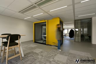 Moderne kantoorruimte met gele geluidscabine, houten stoel met zwart zitgedeelte, grijze tapijttegels en witte wanden.