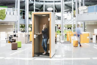 Vrouw in telefooncel met houten frame in modern kantoorinterieur met gele en groene accenten.
