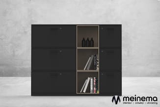 Zwarte kastenwand met drie open vakken in het midden, gevuld met boeken en flessen. Tegen grijze betonnen achtergrond.