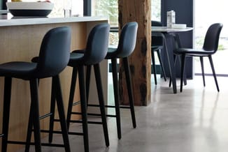 Zwarte barstoel met gevoerde rugleuning aan houten bar in modern kantoor, uitgevoerd met betonnen vloer en raampartijen.