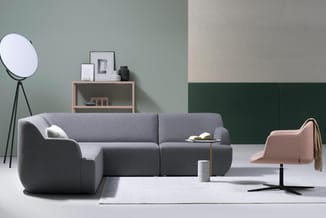 Grijze modulaire bank met roze fauteuil, staande lamp, houten kastje en groene wandpanelen in modern kantoorinterieur.