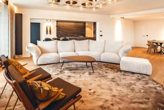 Moderne woonkamer met gekromde grijze zitbank, houten salontafel, bruine leunstoel, houten vloer en beige tapijt.