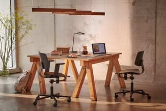 Werkplek met houten tafel op schragen, twee zwarte bureautoelen met wielen, hangende bruine design-lamp, laptop, plant in vaa
