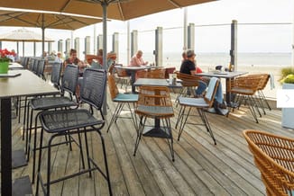 Terras met houten vloer onder parasols, zwarte en rieten stoelen rond tafels, uitzicht op strand en zee, enkele bezoekers zit