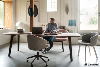 Man aan tafel met laptop, grijze stof bureaustoelen, houten tafelblad, zwarte tafelpoten, lichte kantoorruimte met ramen en d