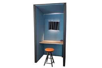 Akoestische belcel met blauwe wanden, houten werkblad, oranje kruk en ventilatierooster aan bovenzijde.