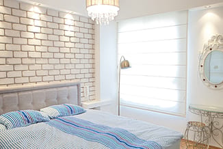 Moderne slaapkamer met grijze bedbank tegen witte bakstenen muur, blauw gestreept beddengoed, hanglamp, spiegel en staande le