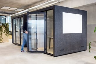 Grijze akoestische belcel met glazen deur en witte whiteboard in moderne kantoorruimte, persoon loopt voorbij.