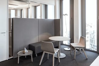 Akoestische werkplek met grijze panelen, beige stoel, kleine ronde tafel en twee witte tafels in modern kantoor met ramen.