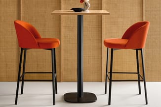 Twee rode barstoeien met zwart metalen onderstel rond een zwart tafeltje met houten blad tegen beige geweven wand.