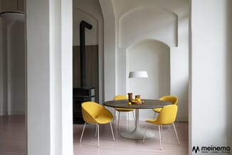 Ronde houten eettafel met wit onderstel, omringd door vier gele stoelen met witte poten in een moderne interieurruimte met wi