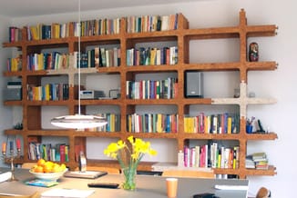 Houten modulaire boekenkasten met vakken tegen witte muur, gevuld met boeken. Houten werktafel met gele bloemen, fruit en kan