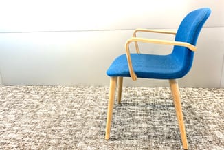Blauwe stoel met houten frame en armleuningen, staand op tapijt tegen witte wand.