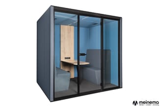 Gesloten belcel met zwart frame, blauwe wanden, houten bureau aan linkerzijde en grijze zitbank rechts.