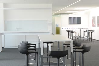 Modern kantoorruimte met witte keukenkast, grijze tafel met zwarte stoelen, bartafel met krukken en TV aan wand.