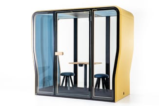 Gele akoestische telefooncel met glazen wanden, zwarte frame, twee blauwe krukken en houten tafelblad tegen witte achtergrond