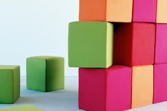 Gekleurde kubussen van schuimstof in groen, rood, roze, oranje en beige, gestapeld en verspreid op grijze ondergrond tegen wi