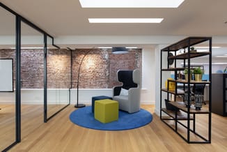 Modern kantoor met grijze fauteuil, geel kubusvormig poef op blauw rond tapijt, zwarte stalen kasten, glazen wanden en bakste