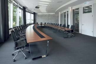 Moderne vergaderruimte met U-vormige houten vergadertafel, zwarte bureaurstoelen, grote ramen, grijs tapijt en spotverlichtin