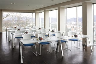 Moderne vergaderruimte met witte tafels, witte stoelen met blauwe zitting, donkere houten vloer en grote ramen met stadsaanzi