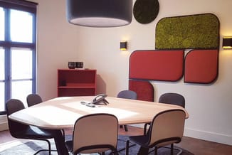 Moderne vergaderruimte met ovale houten tafel, bruine stoelen, rode wandpanelen, groene akoestische elementen, hangende lamp