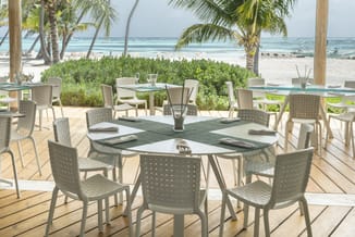 Grijze kunststof stoelen rond witte vergadertafel op houten terras met zee en palmen op achtergrond.