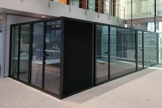 Zwarte glazen belcel met aluminium framewerk in moderne kantoorhal, zijkant en voorkant zichtbaar.