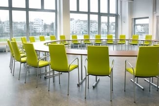Vergadertafel met groene stapelstoelen in moderne kantoorruimte met raampartijen en uitzicht op woongebouw.