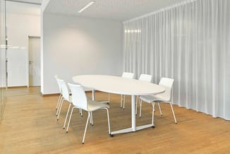 Moderne vergaderruimte met ovale witte vergadertafel, vier witte stoelen, houten vloer en grote raampartij met sheer gordijne