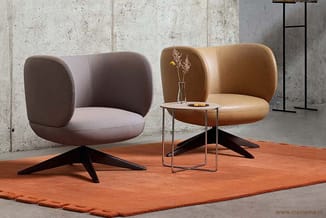 Twee moderne stoelen met zwarte stervoet op oranje tapijt, tussen hen een glazen bijzettafel met bloemen, tegen betonnen muur