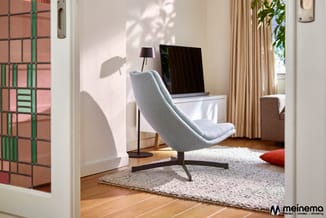 Moderne werkruimte met grijze loungestoel, wit bureau met monitor, zwarte leeslamp en beige gordijnen op houten vloer.