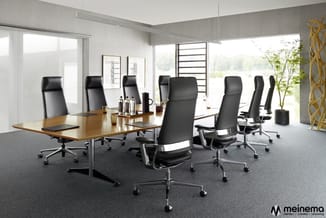 Moderne vergaderruimte met houten vergadertafel, zwarte ergonomische bureaustoelen, grote ramen en grijze vloer.