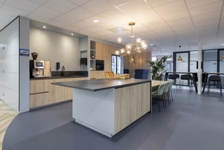 Moderne kantoorkeukentafel met donkergrijs blad en lichte houten zijkanten, omgeven door lichte houten kastenwand en barstoel