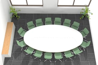 Vergaderruimte met ovale witte tafel en 16 groene kantooorstoelen met wit frame, grijze tegelvloer, planten en raampartijen.