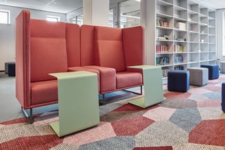 Rode akoestische zitcabine met hoge rug in bibliotheekruimte met groene poefjes, grijze kasten en kleurig tapijt.